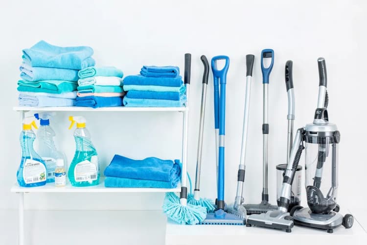 Uma variedade de produtos e equipamentos de limpeza, como borrifadores, panos de microfibra, rodos e aspiradores, organizados em prateleiras ou em um carrinho de limpeza.